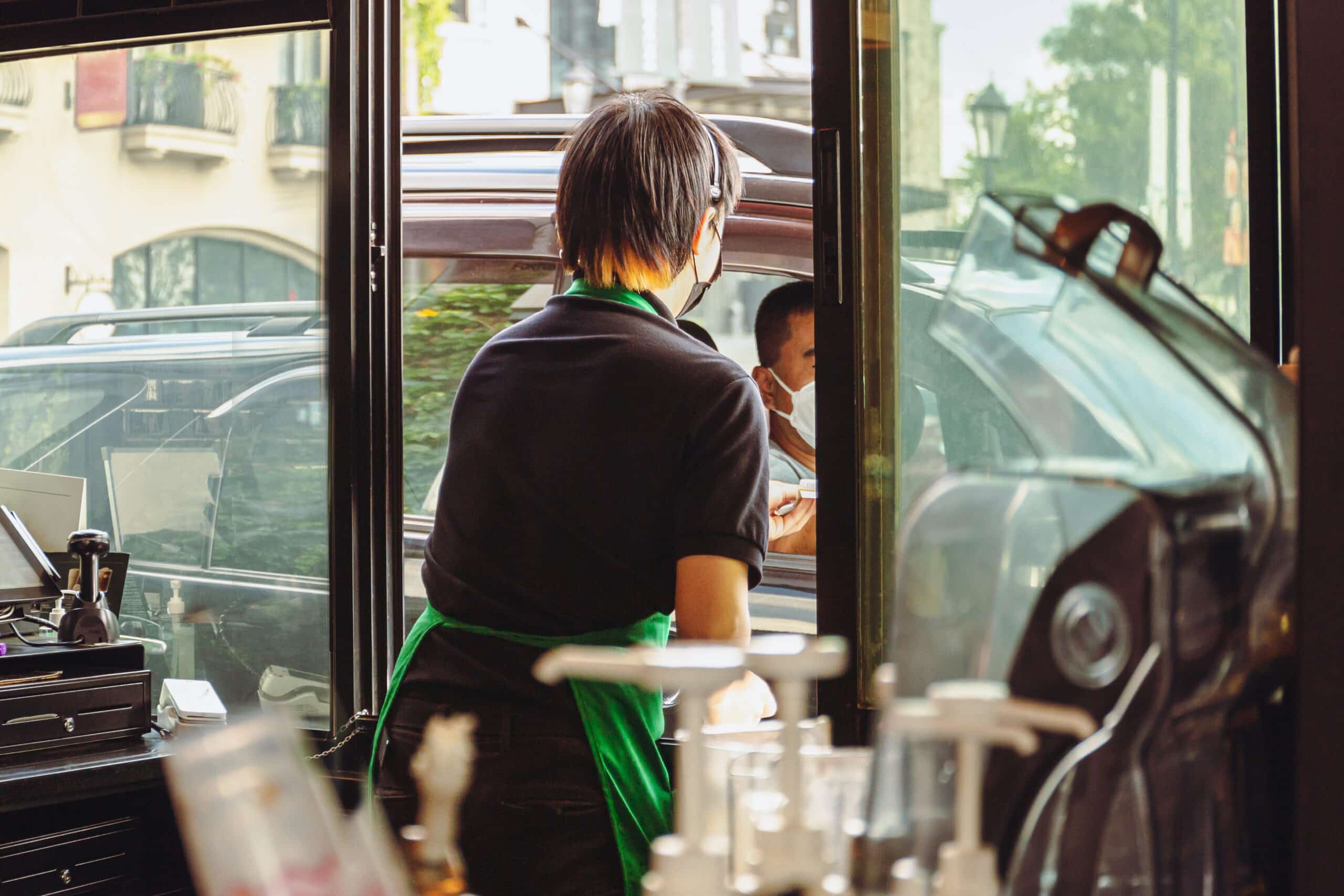 Coffee shop staff with protective mask serving coffee to customer at drive thru.