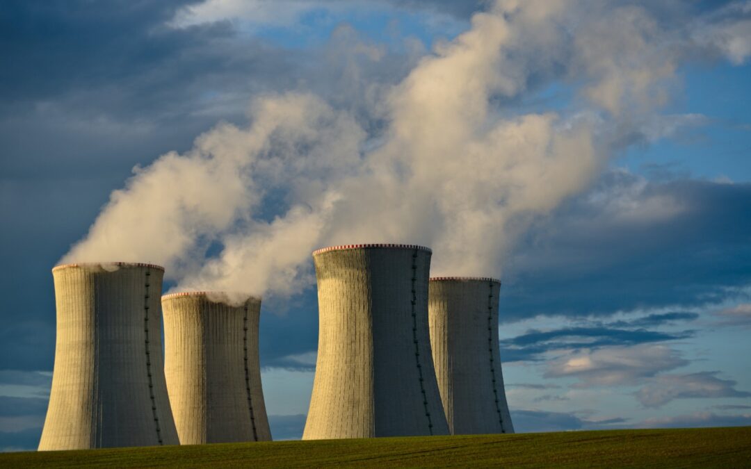 A cooling tower, cylinder, and silo of a nuclear power plant stand tall against a cloudy sky in an outdoor grassy area.